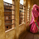 A photograph of a woman wearing a pink sari holding a green-handled broom sweeping in an arched corridor