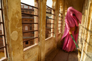 A photograph of a woman wearing a pink sari holding a green-handled broom sweeping in an arched corridor